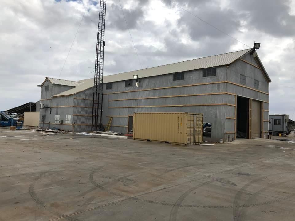 A large gray industrial warehouse building with a metal tower and a tan shipping container on a concrete lot.