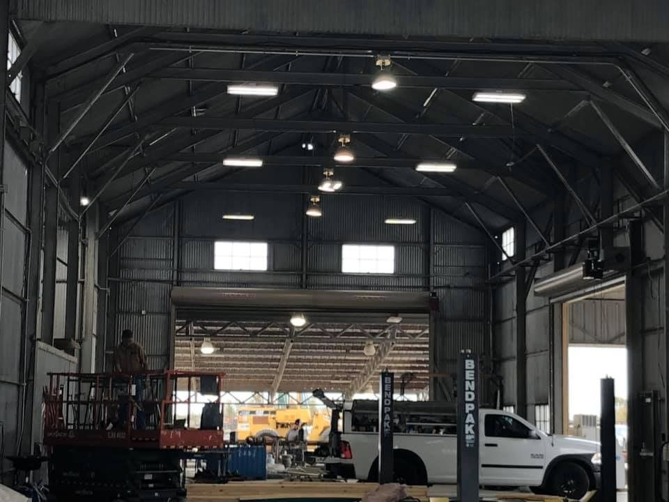 An interior view of a large, high-ceilinged industrial workshop with a white pickup truck and a red scissor lift.