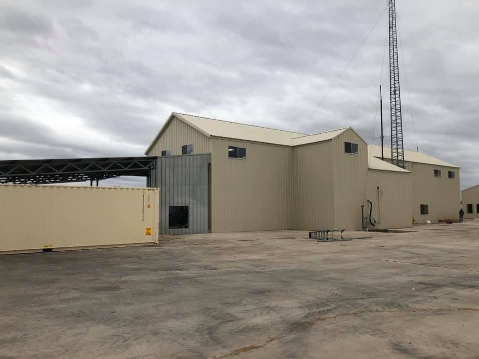 A tan metal building with a shipping container parked in front, under a cloudy sky.