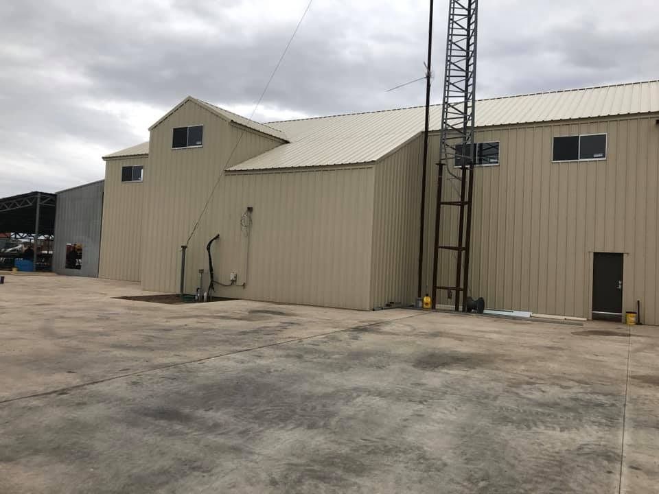 A beige, industrial metal building with a metal tower and an asphalt parking lot under a cloudy sky.