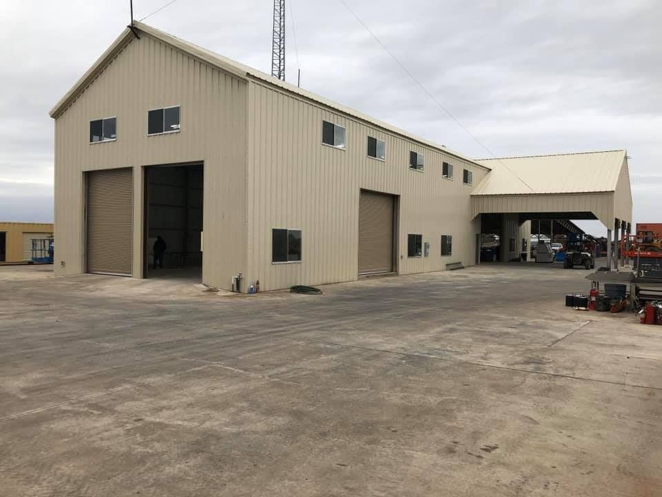 A large, beige metal industrial building with roll-up doors and windows under a cloudy sky.