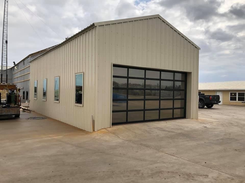 A tan metal warehouse building with a large glass sectional garage door and four vertical windows on a concrete lot.