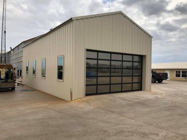 A tan metal warehouse building with a large glass sectional garage door and four vertical windows on a concrete lot.