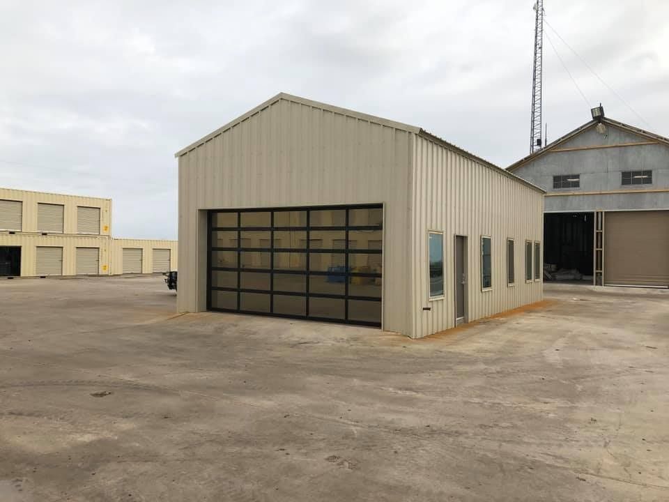A tan metal commercial building with a large glass sectional garage door, surrounded by a paved lot on a cloudy day.