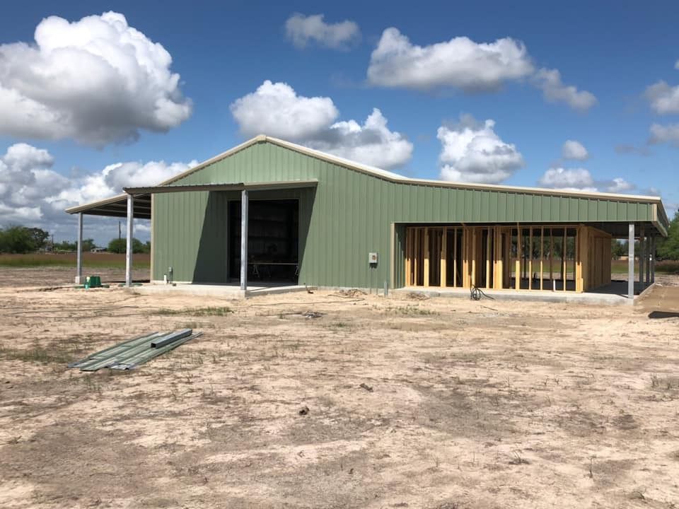 A partially constructed green metal building with exposed wooden framing on the right side against a blue, cloudy sky.