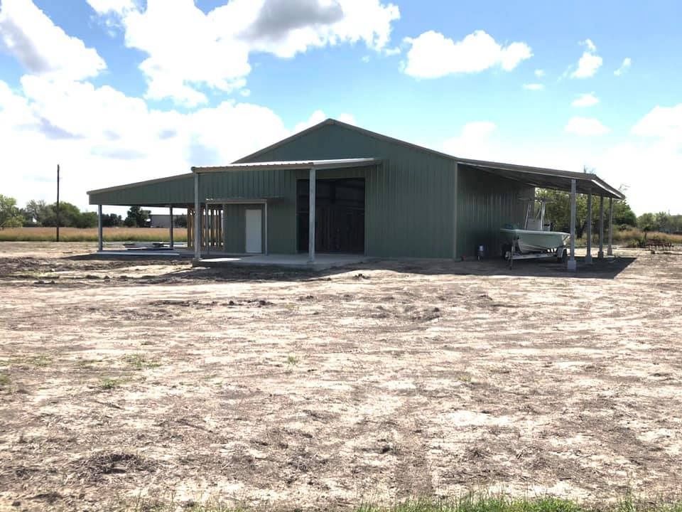 A green metal building with a central entrance and open-sided lean-tos stands on a flat, dirt lot under a blue sky.