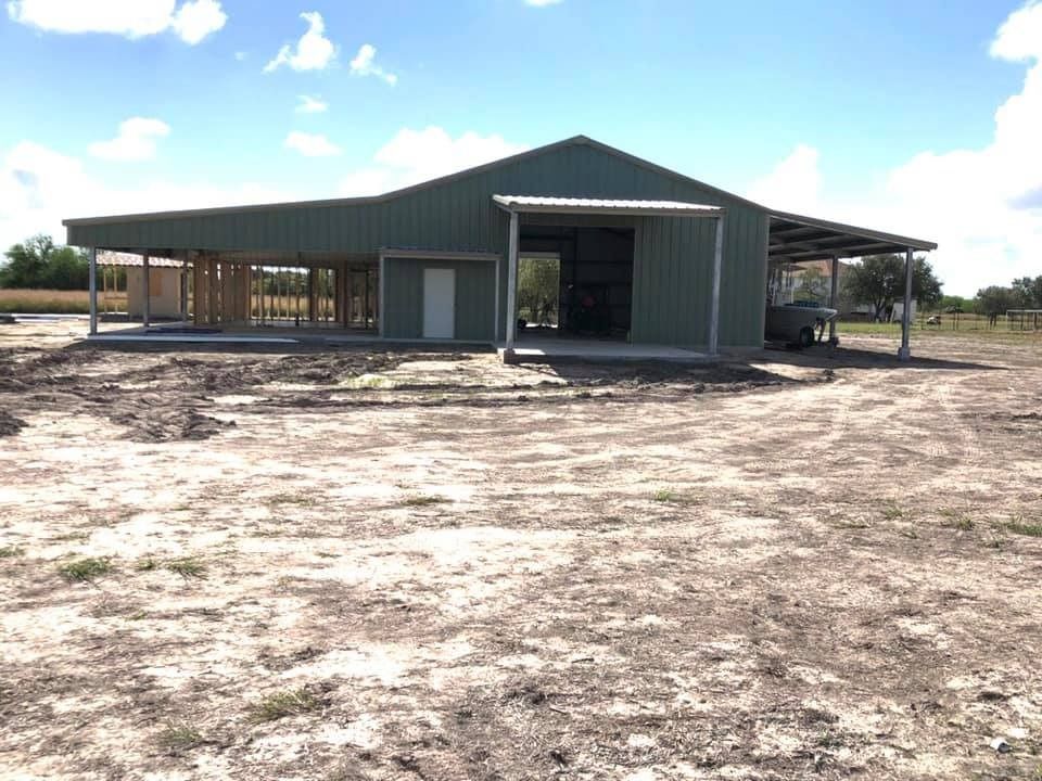 A green metal barn with an open carport area under construction on a dirt lot under a sunny, blue sky.
