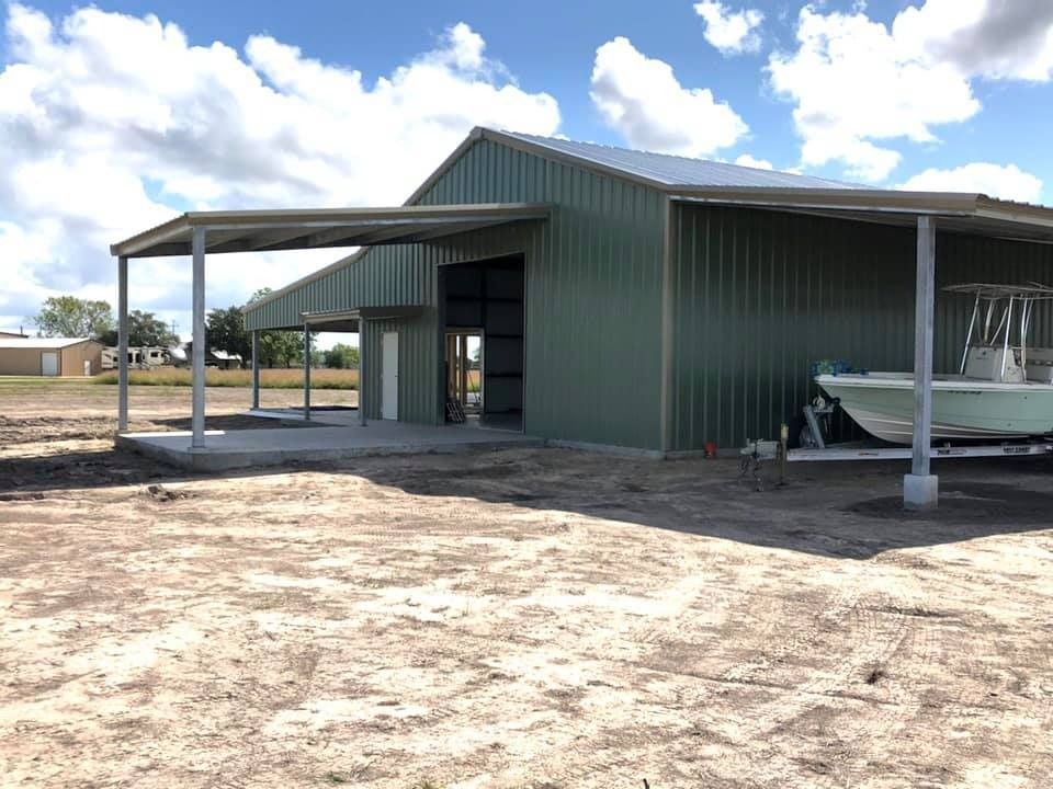 A green metal pole barn structure with a large open doorway and side covered parking sheltering a white boat on a trailer.