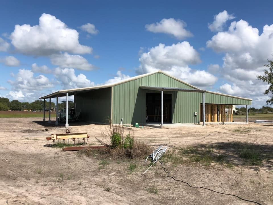 A green, metal-sided barn or workshop under construction on a sunny, grassy lot under a bright, cloudy sky.