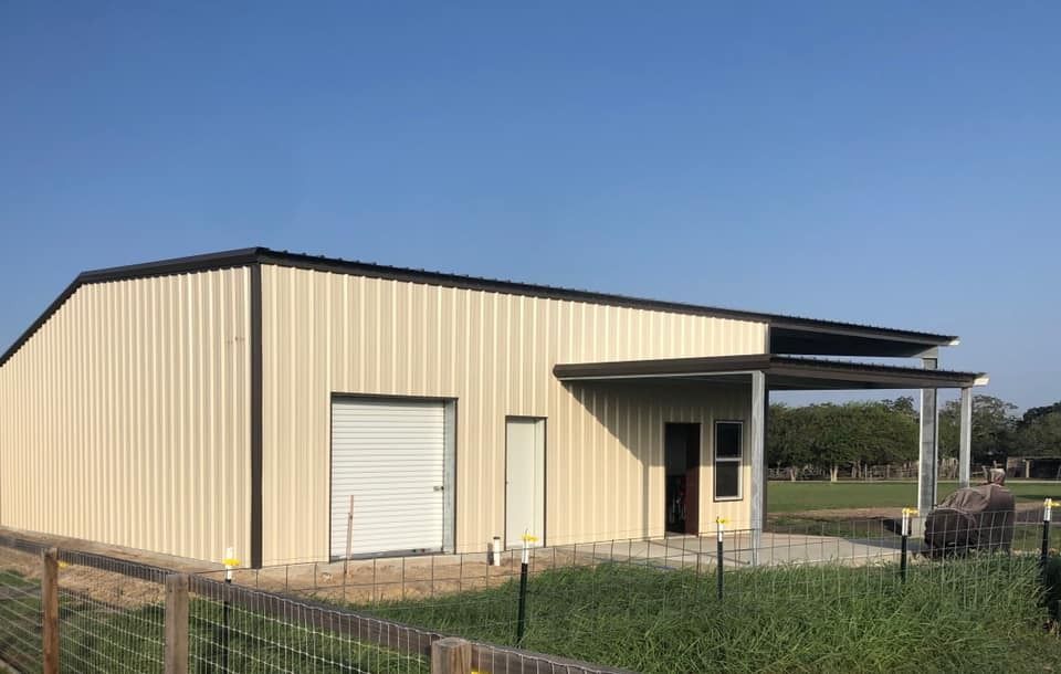 A beige metal workshop with a roll-up door, a side door, and a covered front porch, situated on a grassy lot.