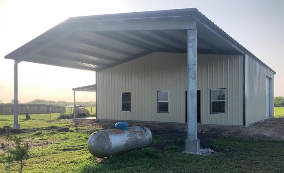 A light-colored metal barn with a wide carport roof extension, standing in a rural field with a large propane tank nearby.