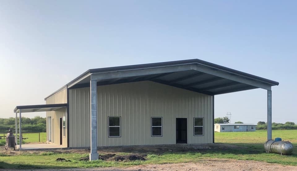 A tan metal building with a large covered porch area set in a grassy field under a clear blue sky.