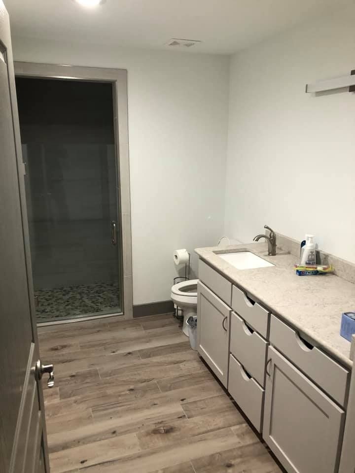 A modern bathroom featuring light gray cabinets, a white countertop, a toilet, and a walk-in glass shower with wood floors.