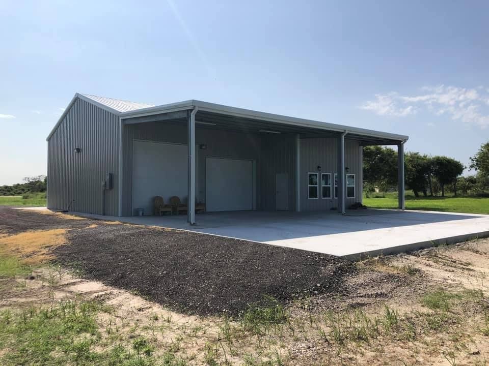 A gray metal workshop with a large open-air covered porch and concrete slab, set in a sunny, rural, grassy landscape.