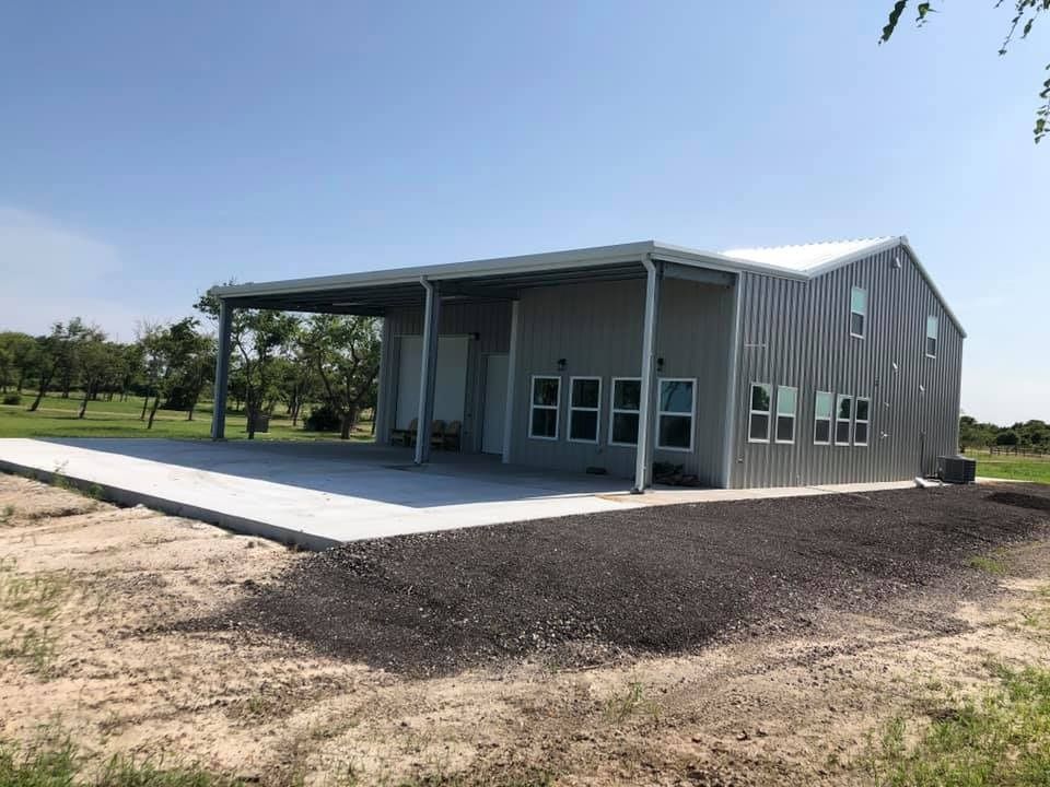 A modern gray metal building with a wide covered patio, set in a rural landscape with trees under a clear blue sky.