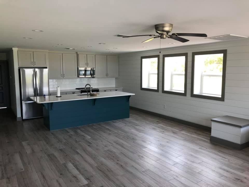 A modern kitchen featuring a teal island, stainless steel refrigerator, light grey cabinets, and wood-look flooring.