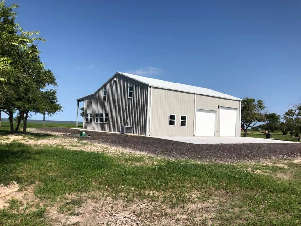 A modern, light-colored metal building with two garage doors sits on a gravel lot under a clear blue sky.