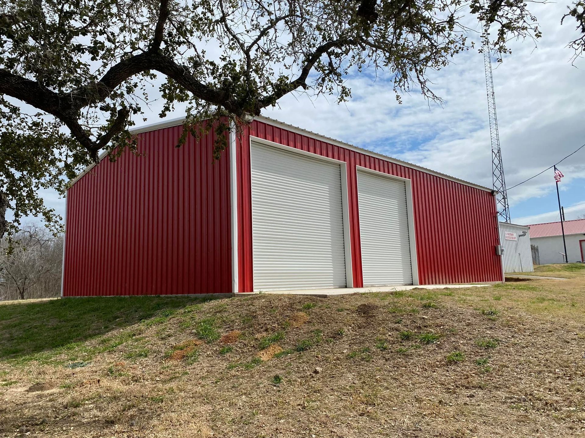 A bright red metal storage building with two white roll-up doors, set on a grassy hill under a cloudy sky.