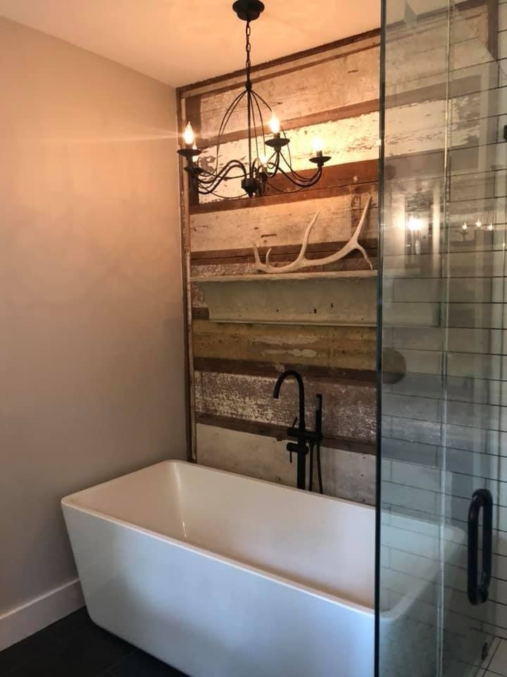 Modern white freestanding tub in a bathroom with a rustic reclaimed wood feature wall, chandelier, and glass shower.