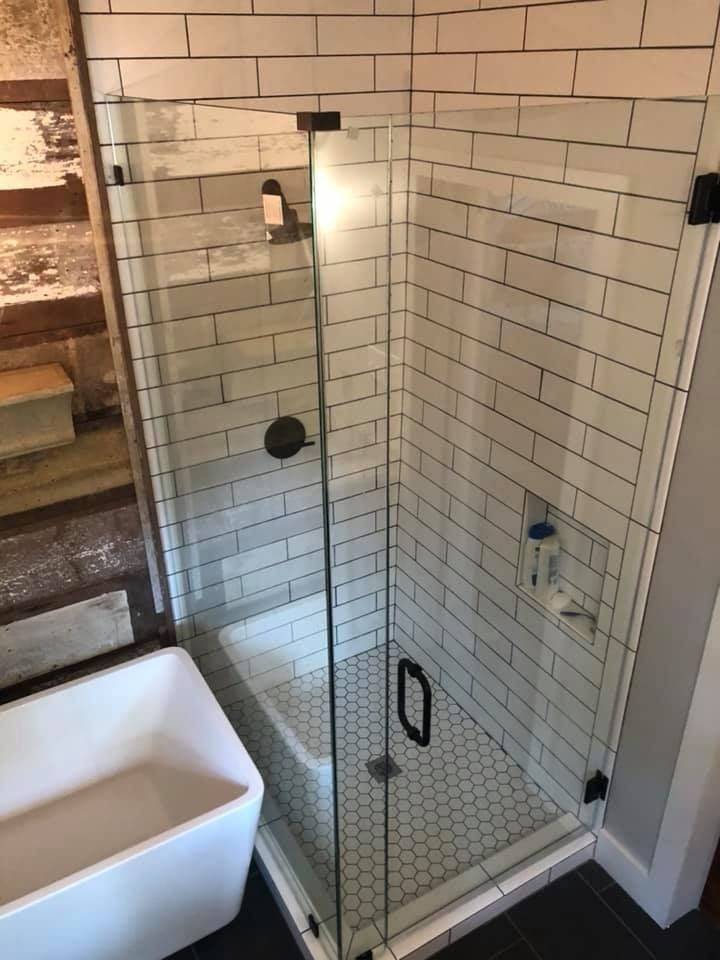 A walk-in shower with white subway tile walls, a hexagon tile floor, a glass door, and black hardware next to a bathtub.