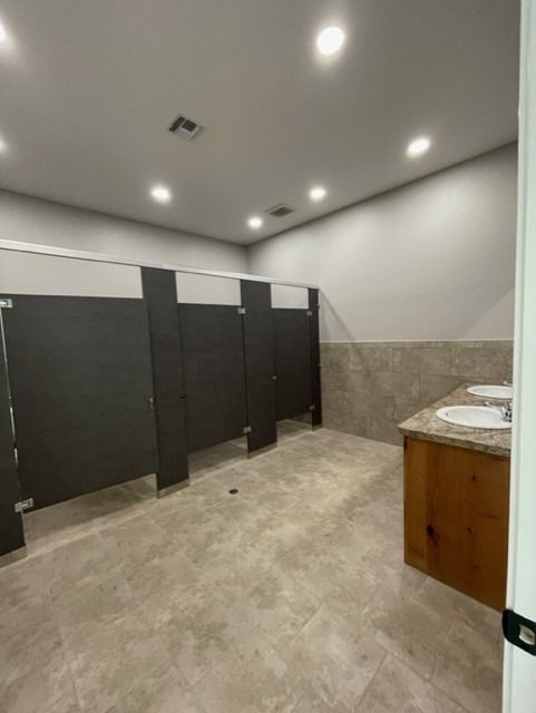 A public restroom featuring dark gray stall partitions, tan tiled walls, and a wooden vanity with two sinks.