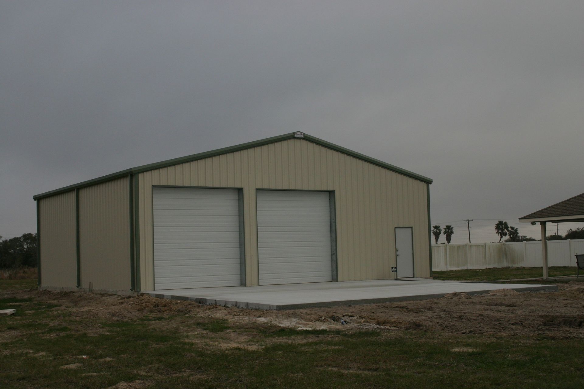A beige metal garage with two white roll-up doors and one side entry door stands on a concrete slab in a grassy field.