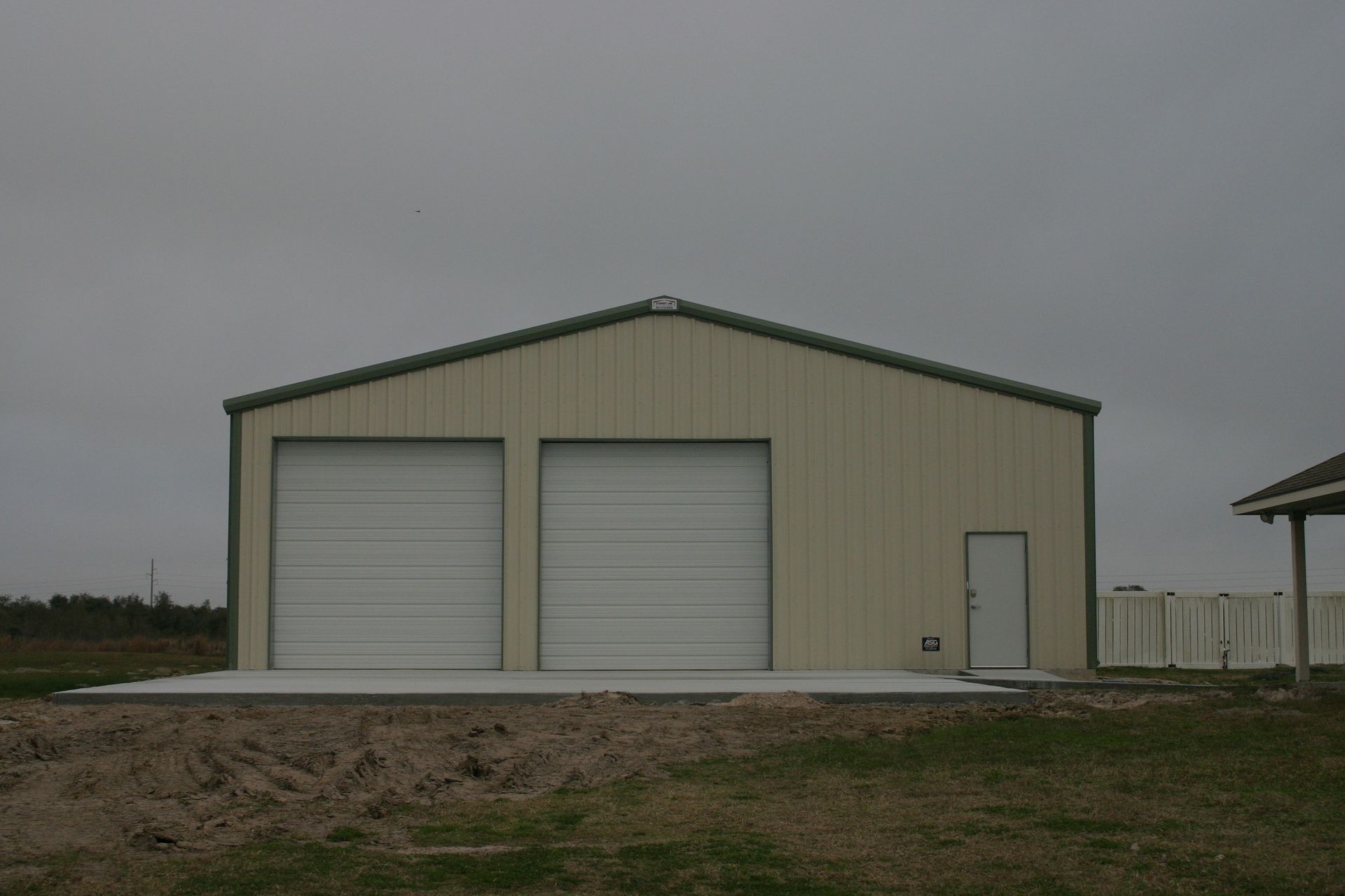 A tan metal garage building with two white roll-up doors and a side entry door set on a concrete pad under a gray sky.
