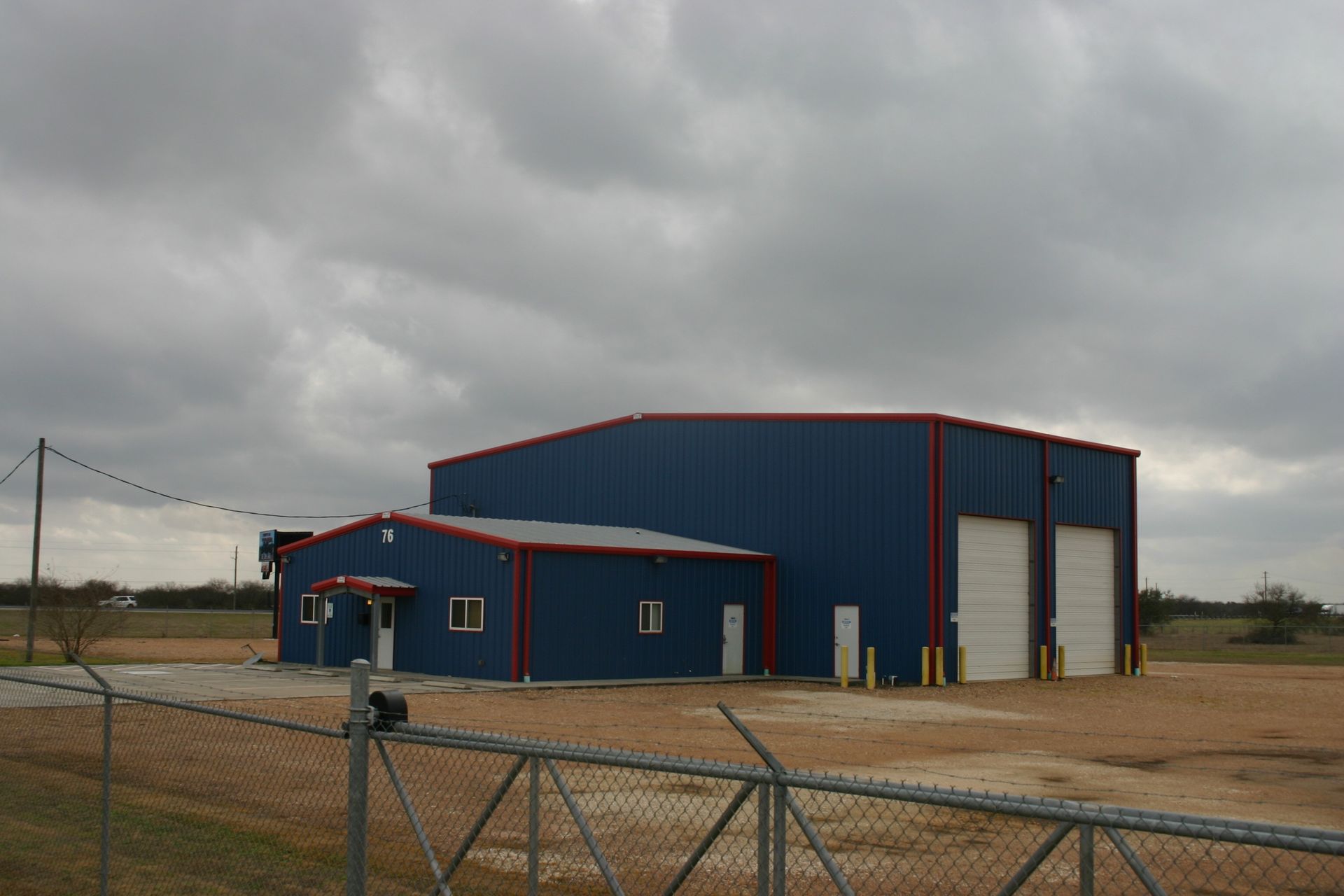 A blue metal warehouse building with a smaller attached office, red trim, and two garage doors under a cloudy sky.