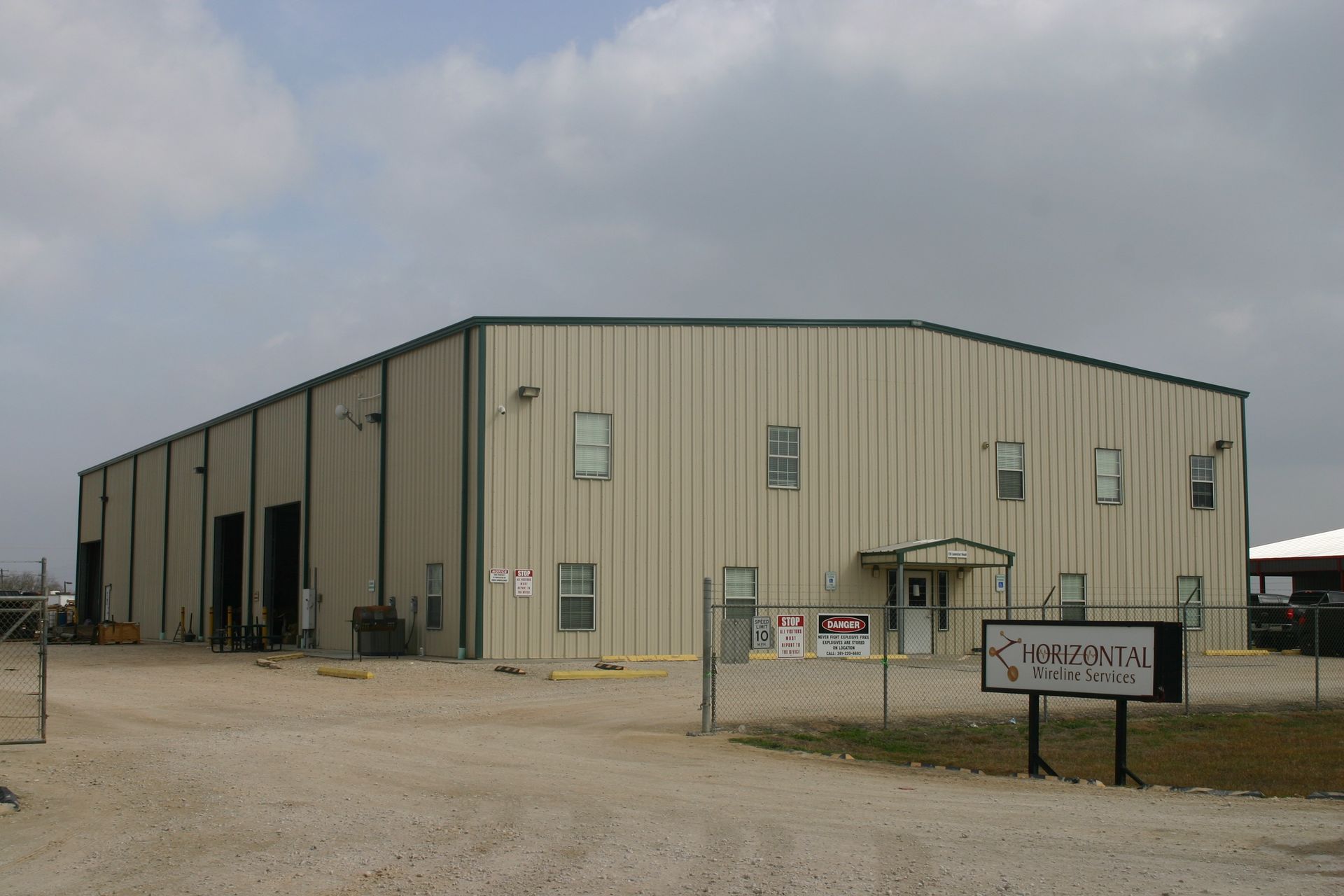 A tan, industrial warehouse building with a metal exterior, multiple windows, and a gated gravel entrance.