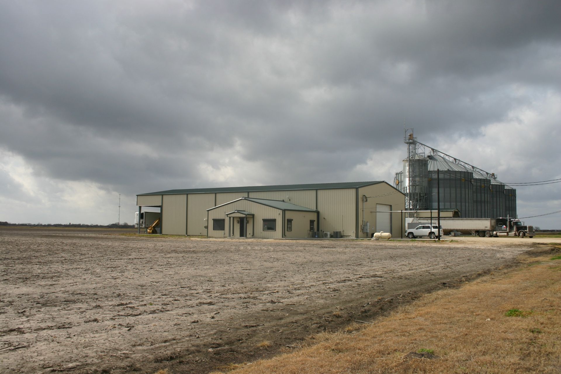 A large metal agricultural building with grain silos stands in a flat, plowed field under a cloudy, overcast sky.