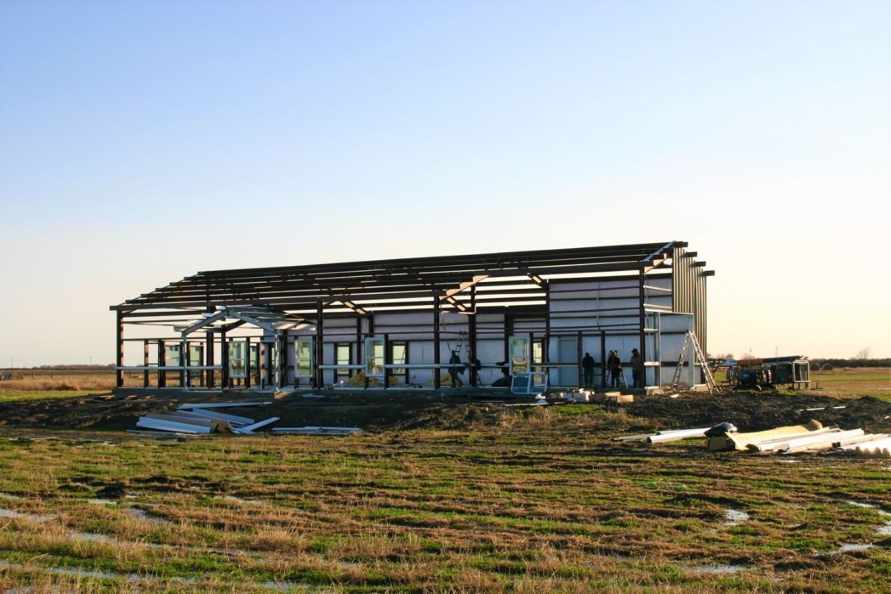 A partially constructed metal-frame building stands in a flat, grassy field under a clear blue sky.