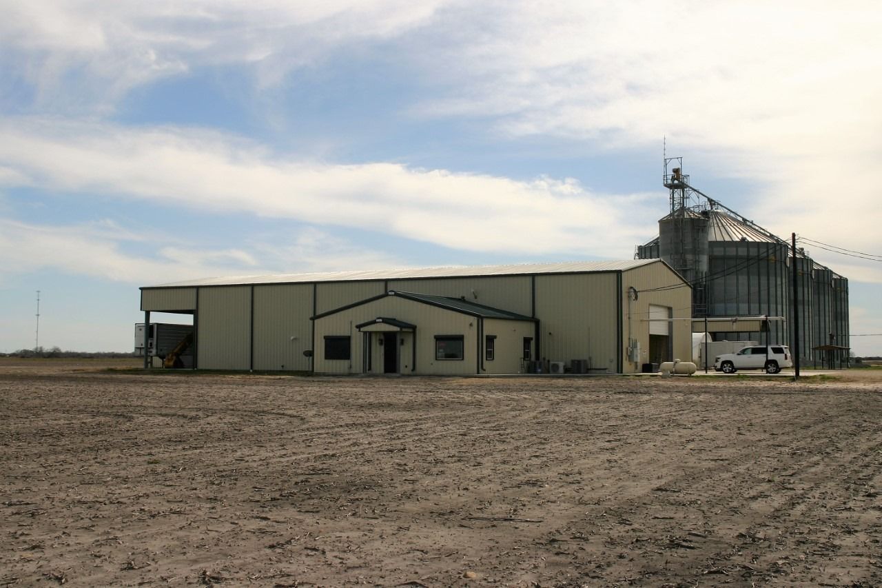A tan commercial building with a grain silo in a large, empty field under a cloudy sky.