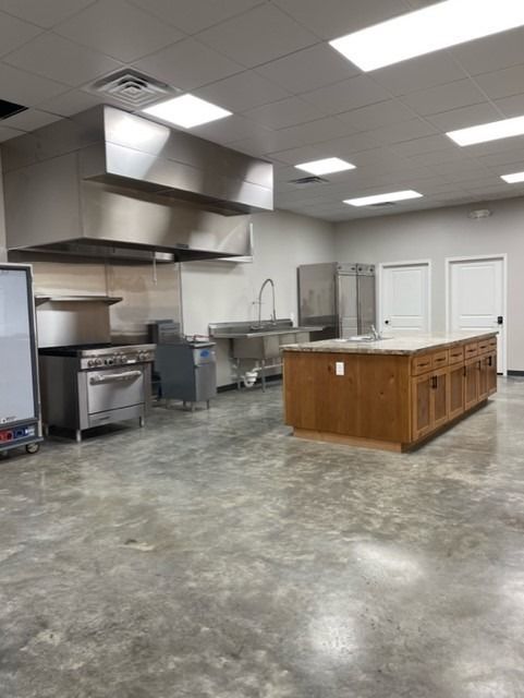 A clean commercial kitchen with a stainless steel range under a hood, a central wooden island, and a sink on the wall.