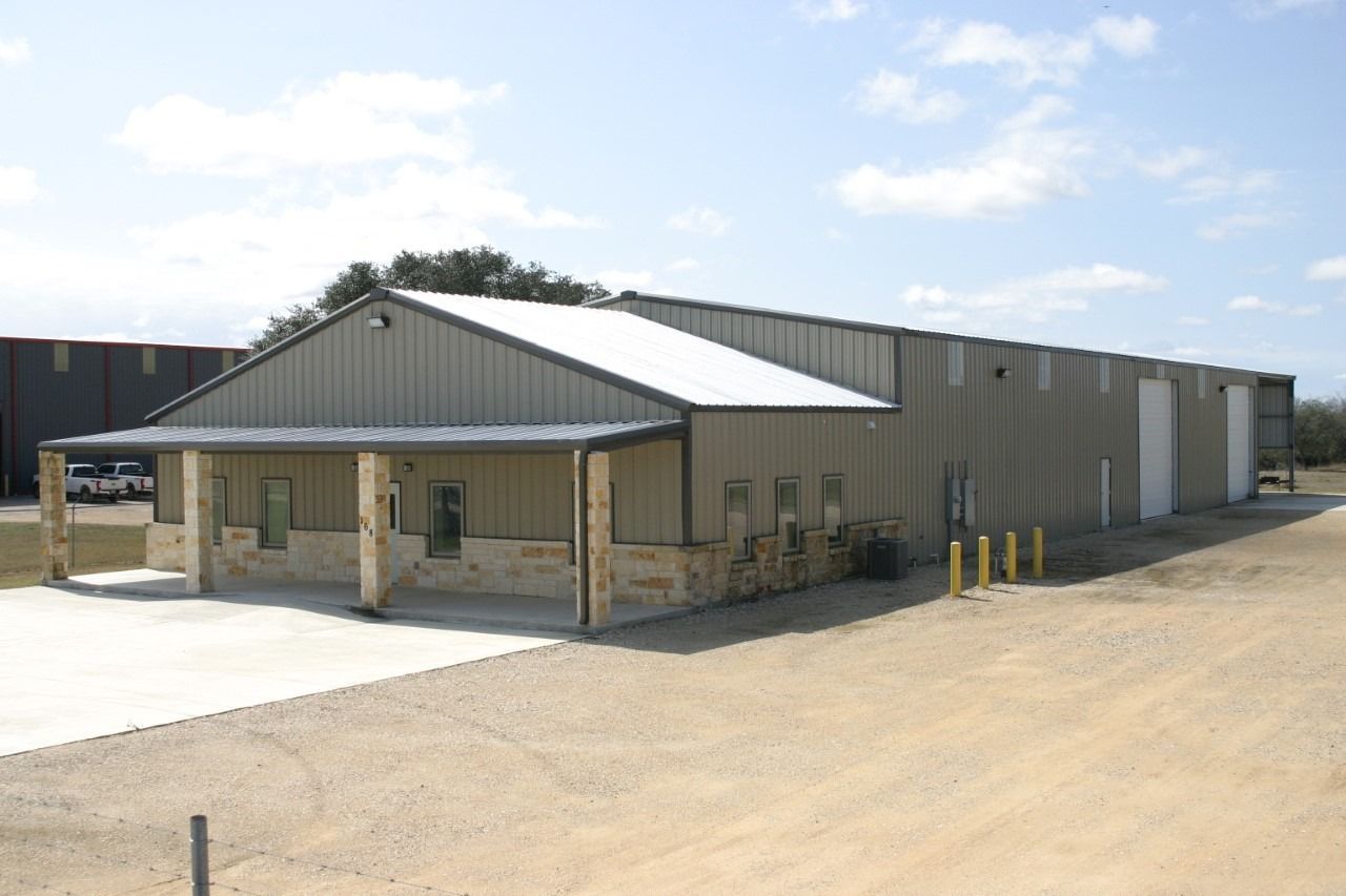 Tan metal warehouse building with a covered stone-pillar entryway and a concrete pad in a rural, sunny setting.