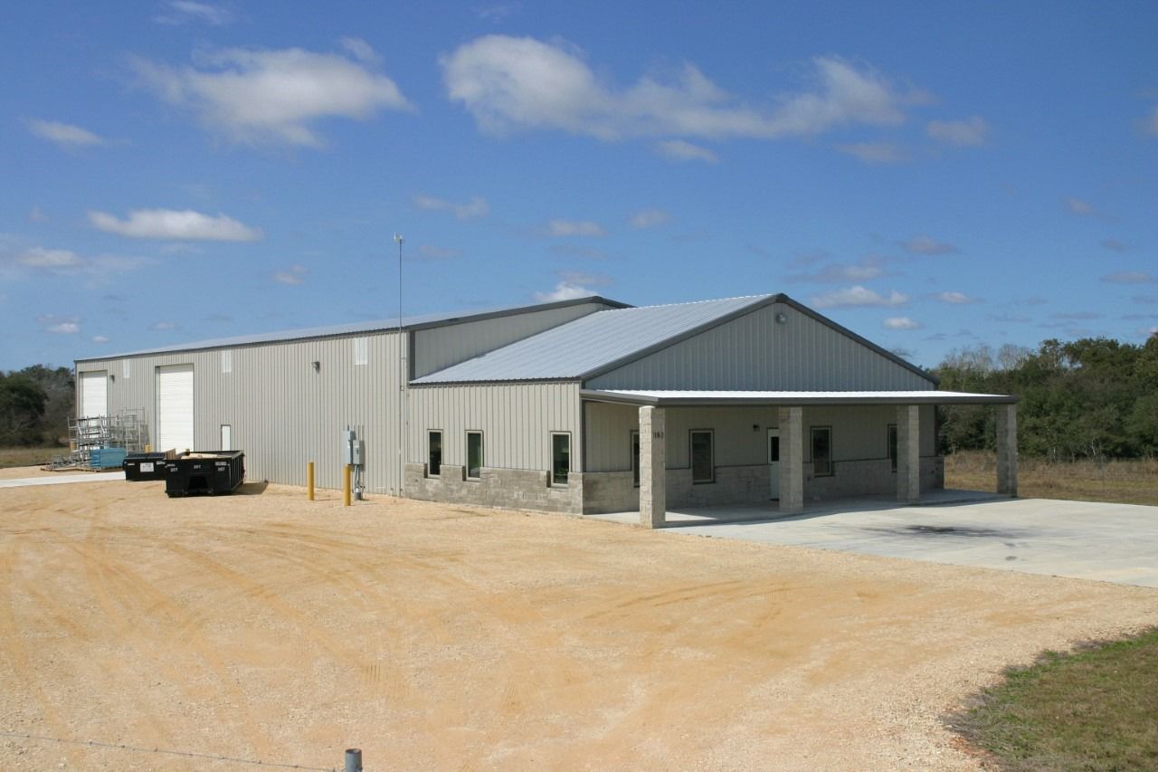 A large, gray metal warehouse building with a covered front porch and a gravel parking lot under a blue sky.