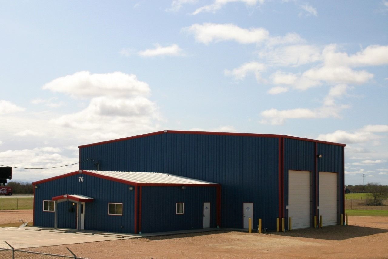 Blue metal building with a white roof, an attached office section, and two large garage doors under a blue, cloudy sky.