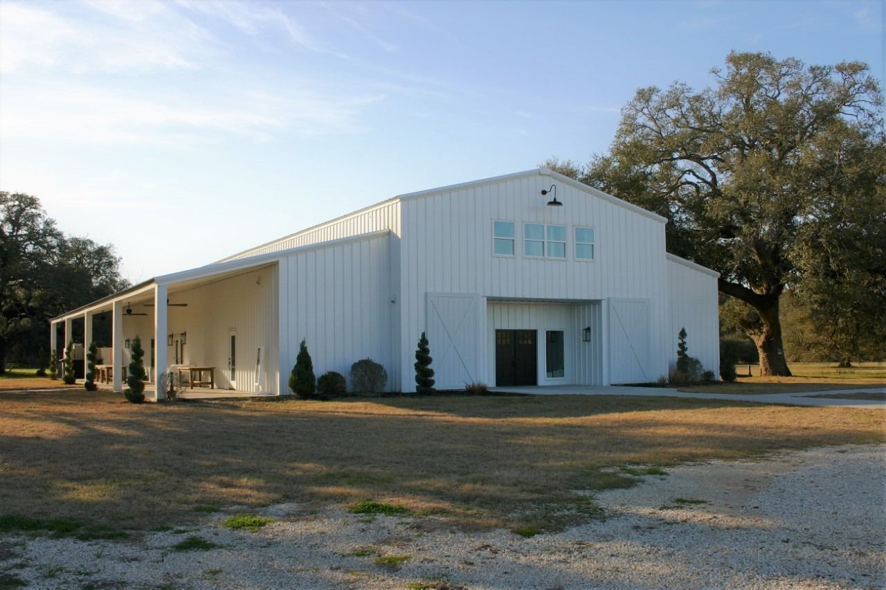 A white modern barn-style building with a covered side porch, standing in a field with large trees under a clear sky.