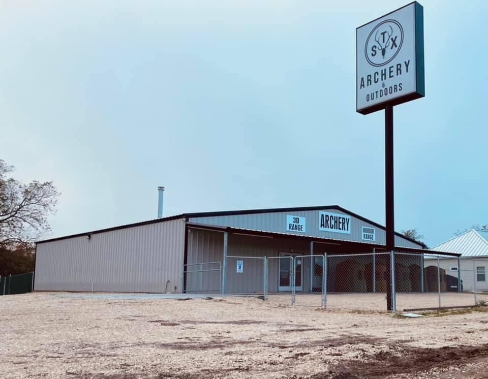 A light-colored metal building with an archery range sign, set on a gravel lot under a pale sky.