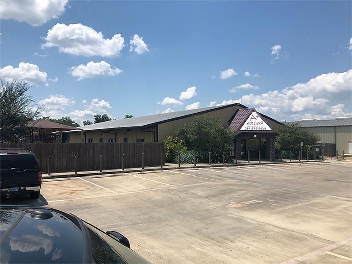 A tan, one-story commercial building with a dark roof and parking lot under a bright, partly cloudy blue sky.