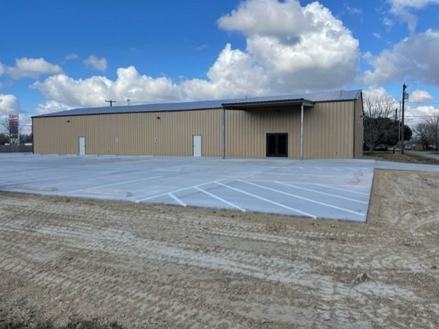 A tan metal warehouse building with a paved parking lot in front under a blue sky with white clouds.