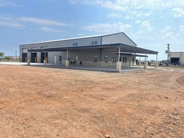 A large, light-gray metal commercial building with a covered exterior walkway on a dirt lot under a blue sky.