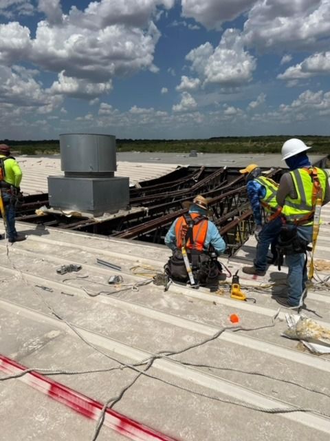 Workers in safety harnesses and hard hats install equipment on a large, flat rooftop under a blue sky.