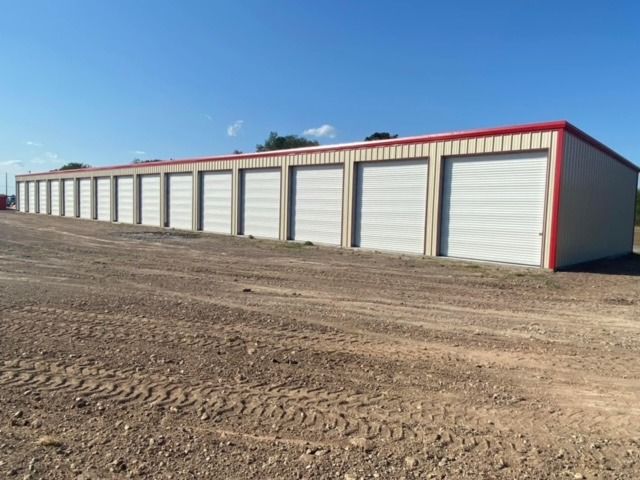 A long, beige metal self-storage facility with white roll-up doors and red trim, set against a clear sky.