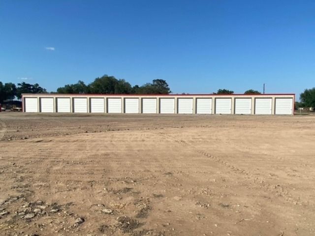 A long, single-story row of storage units with white roll-up doors against a clear blue sky and a dirt lot.