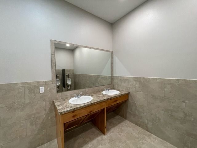 A restroom with a wooden vanity featuring two sinks, a large mirror, and grey tiled walls.
