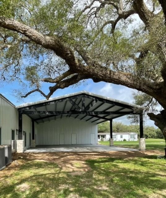 A white metal-framed outdoor pavilion with a concrete floor sits beneath a large, spreading oak tree on a sunny day.