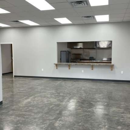 A bright, empty commercial space with polished gray concrete floors, pale walls, and a service window to a kitchen area.