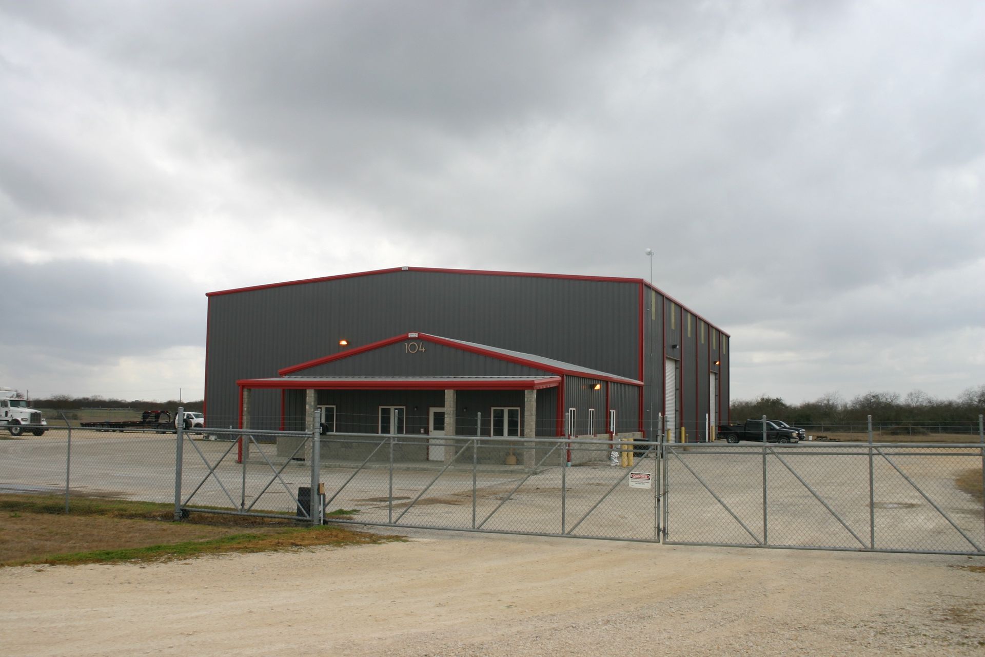 A grey metal warehouse building with a red-trimmed front office entrance, seen behind a chain-link fence under a cloudy sky.