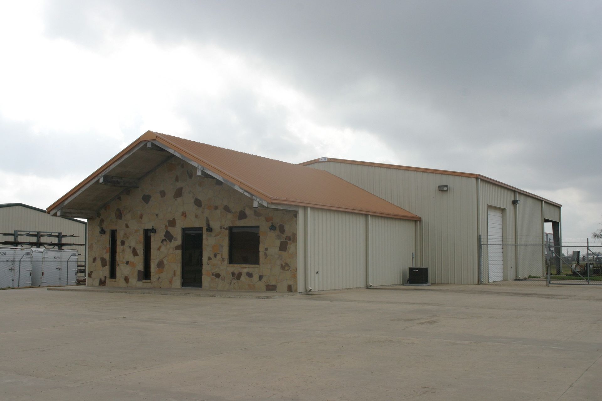 A one-story building with a brown metal roof, a stone-front facade, and tan corrugated metal siding under a cloudy sky.