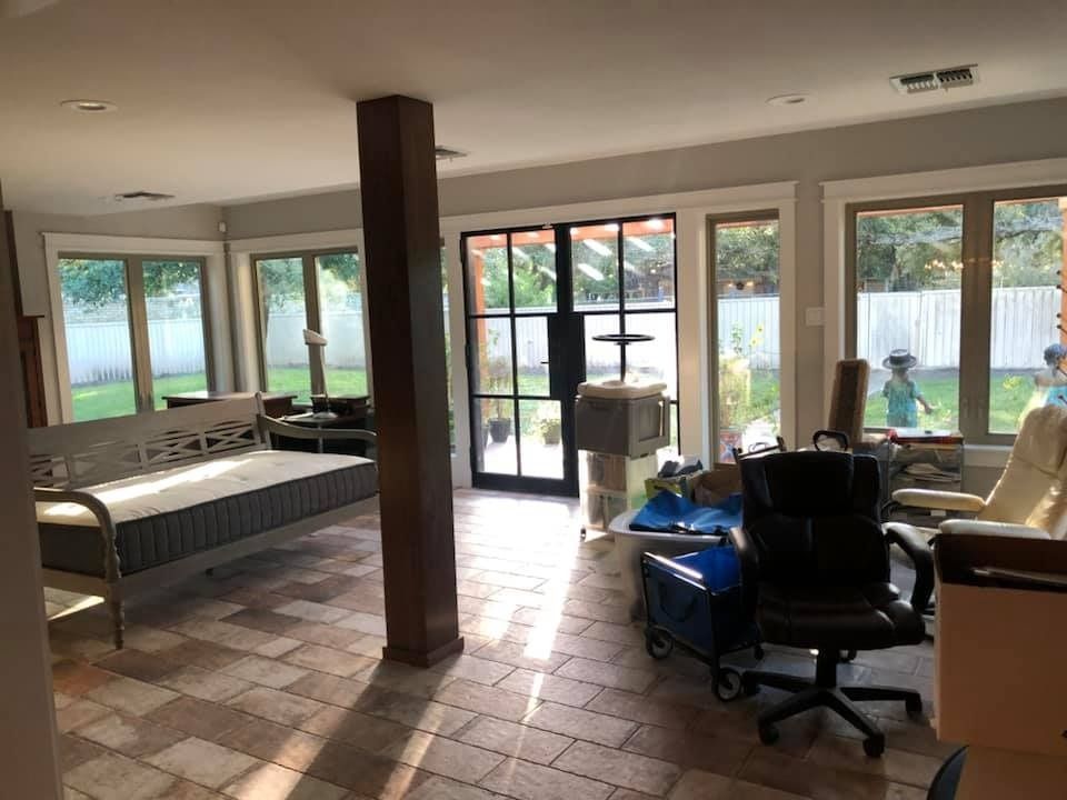 A sunlit room with stone floors, a daybed, a structural column, office chairs, and glass doors looking out onto a yard.